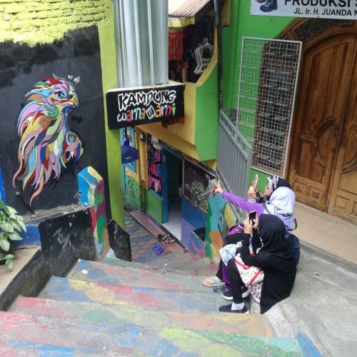 Malang, Indonesien: Zwei Mädchen mit Kopftuch sitzen auf einer bunt bemalten Treppe und fotografieren ein Wandbild ab.